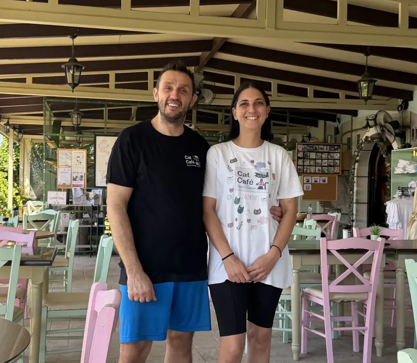 Sakis and Dimitra standing among the tables of their garden, both in Cat Café shirts